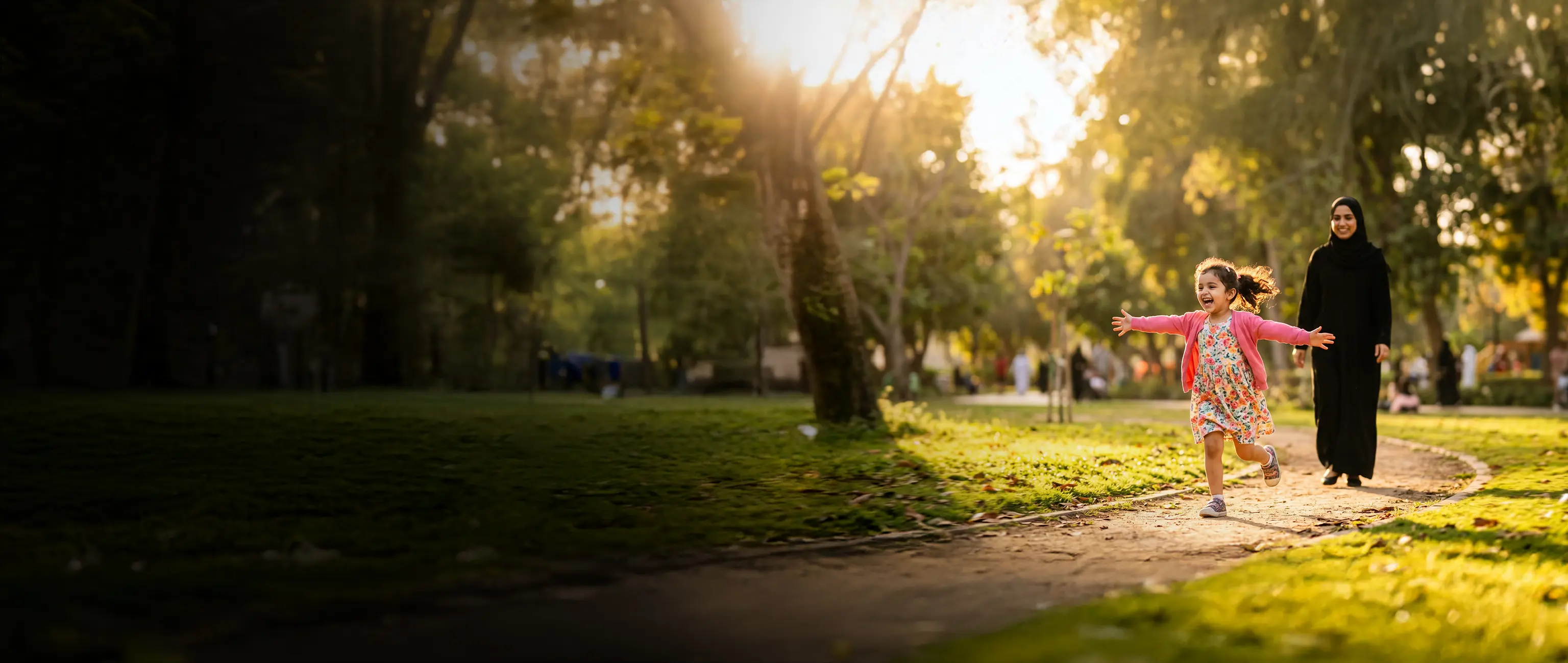 A mother watching her young daughter run happily in a sunny park, symbolizing a secure financial future with kids growing up savings plans from National Bonds.