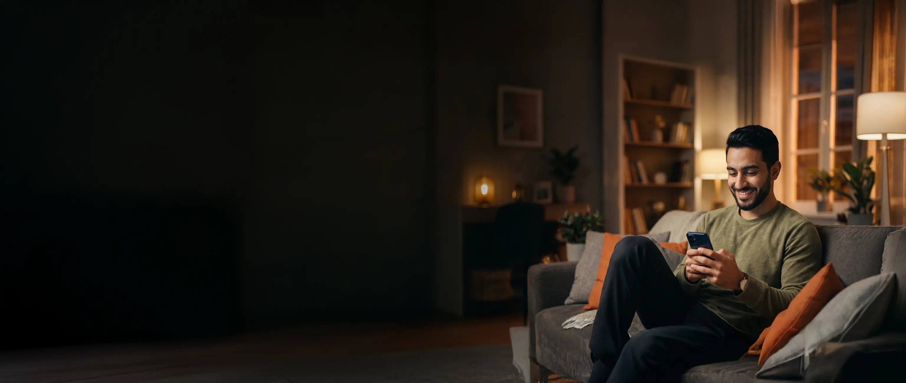 A smiling man relaxing on a couch at home while easily managing his saving plans on his smartphone, representing financial peace of mind and smart saving with National Bonds.